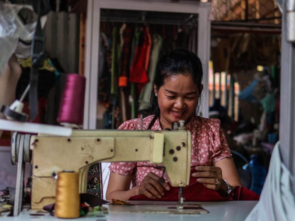 A smiling Cambodian woman named Bunnak sewing traditional clothing at Orussey Market.