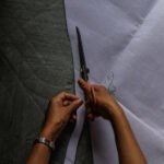 A tailor's hands using large scissors to cut white fabric for a traditional wedding dress.