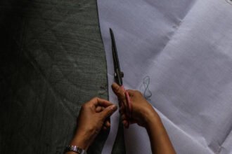 A tailor's hands using large scissors to cut white fabric for a traditional wedding dress.